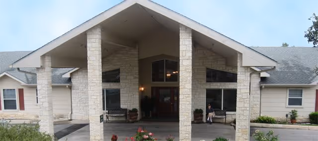 Front entrance of a senior living building with a covered porte-cochère supported by stone columns, double glass doors, benches, and landscaping.