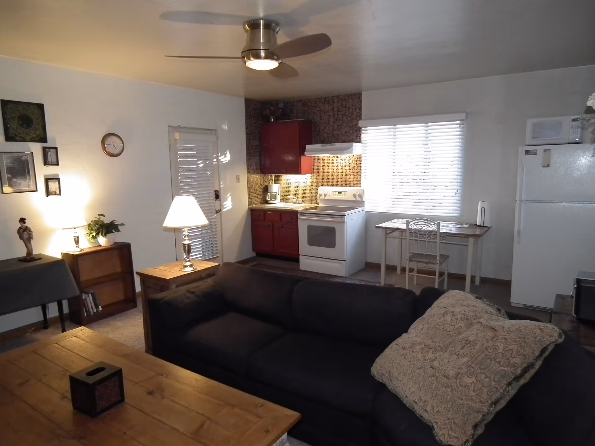 Interior view of a cozy living space featuring a dark sofa with a patterned cushion, a wooden coffee table, a small dining table with a chair near a window with blinds, and a compact kitchen area with red cabinets, a white stove, and a refrigerator. The room is softly lit by table lamps and a ceiling fan with a light.