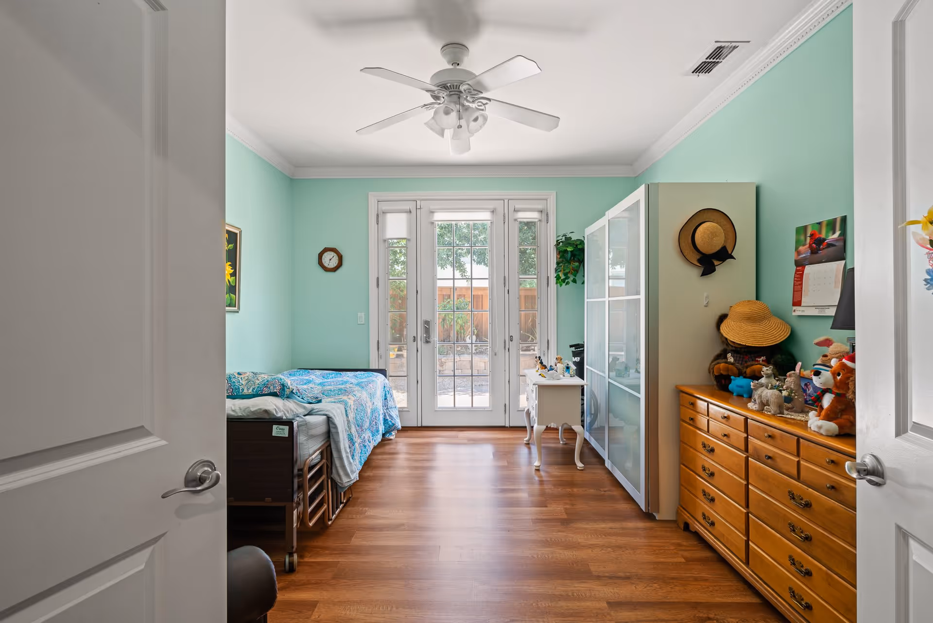 A bedroom with light green walls, a wooden floor, and a ceiling fan. There is a bed with a blue patterned bedspread on the left side, a white cabinet with glass doors, a small white table, and a wooden dresser on the right side. The dresser has various stuffed animals and hats on top. At the far end, there are glass doors leading to an outdoor area with a wooden fence and greenery.