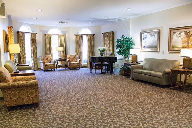 A spacious living room area with patterned carpet, several armchairs and sofas arranged around the room, a black grand piano, side tables with lamps, large windows with curtains, and framed artwork on the walls.