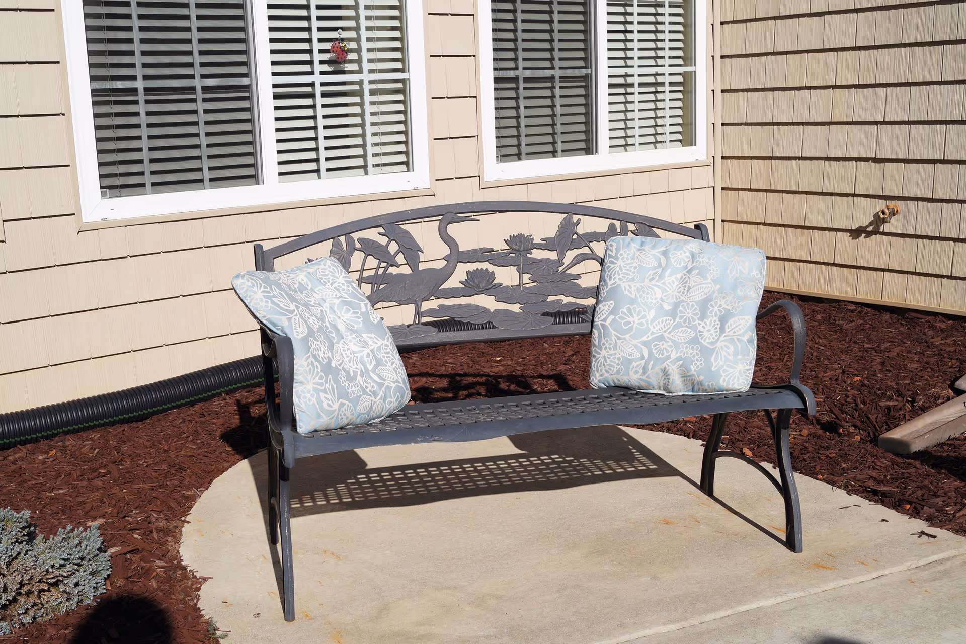 A metal outdoor bench with two patterned cushions on a concrete pad in front of a beige building with windows.