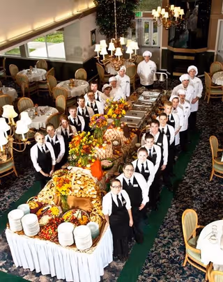 A line of waitstaff and chefs pose beside an elaborate buffet spread in a formal dining room.