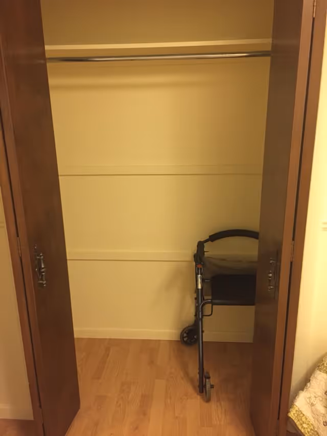 An empty closet with wooden bi-fold doors open, showing a metal walker with a seat inside. The closet has a wooden floor and a hanging rod but no clothes.
