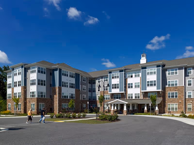 Exterior view of a multi-story senior living facility building under a clear blue sky with a few clouds. The building features a combination of brick and siding with many windows. Two people are walking on the paved driveway in front of the entrance, which has a covered porch supported by columns. Landscaping with small trees and shrubs surrounds the driveway.
