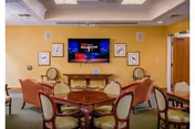 A common area in a senior living facility with several wooden tables and cushioned chairs arranged for group seating. A flat-screen TV is mounted on a yellow wall above a wooden console table, with framed artwork on either side. The room has a carpeted floor and a ceiling with recessed lighting.