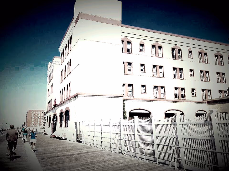Boardwalk alongside a multi-story pale building with a white fence and people walking and biking.