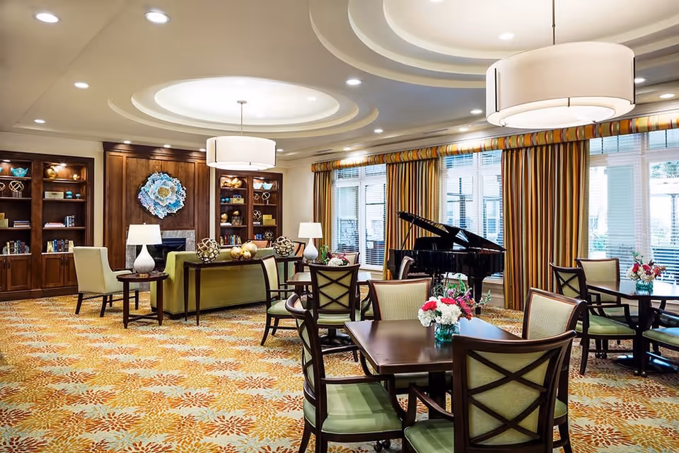 Well-lit senior living common room with tables and chairs, a grand piano, sofas, bookshelves, and large windows with striped curtains.