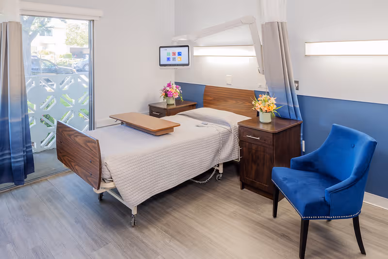 A neatly made hospital-style bed with a wooden headboard and footboard in a bright room. The bed has a beige quilted blanket and a wooden overbed table. On either side of the bed are dark wooden nightstands with colorful flower arrangements. A blue upholstered chair is positioned near the bed. The room has light-colored walls with a blue accent wall and a large window with blue and white curtains, letting in natural light. A mounted screen is visible above the bed.