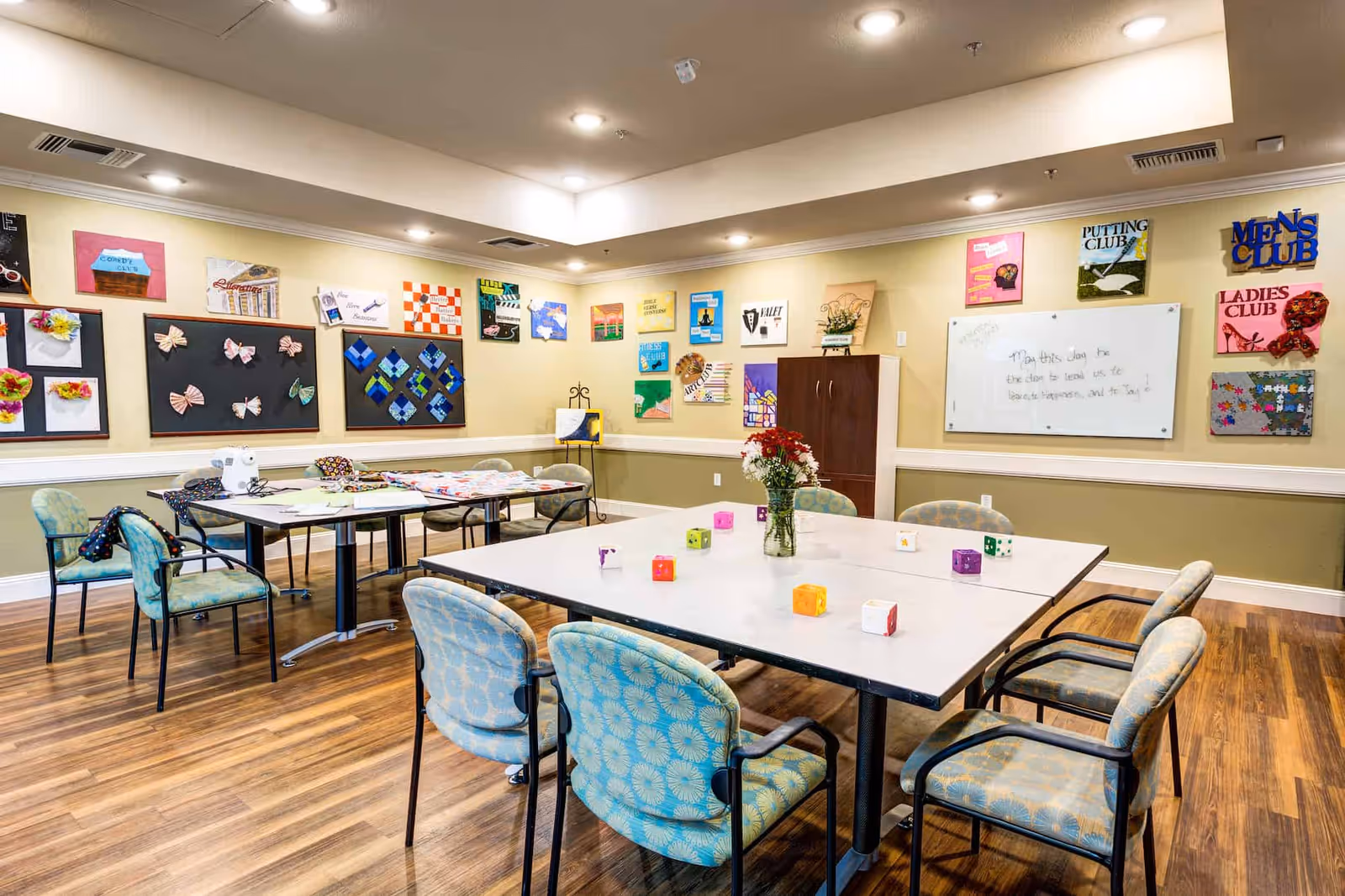 A well-lit activity room with tables and chairs arranged for group activities. The walls are decorated with colorful artwork and club signs such as Men's Club, Ladies Club, and Putting Club. A whiteboard with a motivational message is also visible. The room has a wooden floor and a cabinet in the corner.