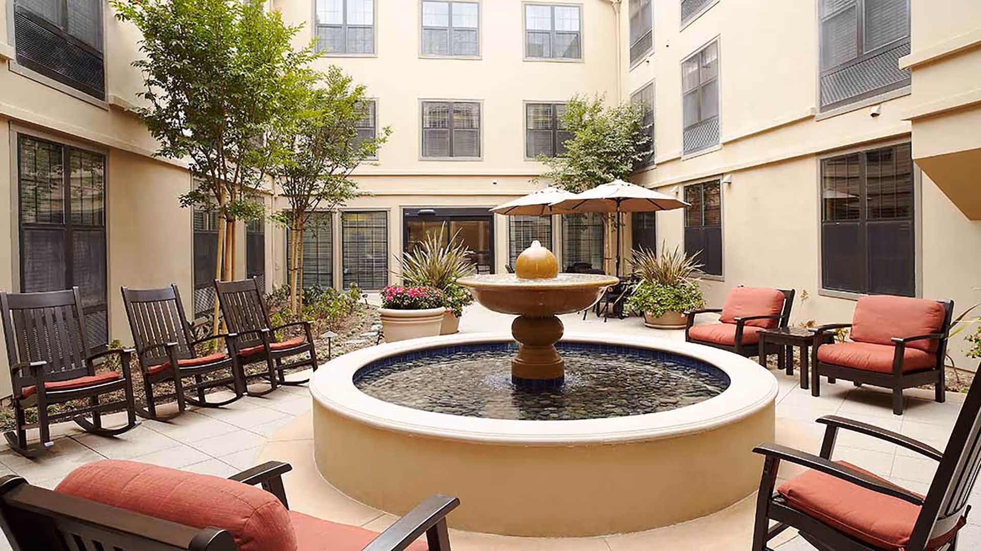 A sunlit courtyard with a central round fountain surrounded by cushioned chairs, rocking chairs, potted plants and umbrellas inside a multi-story building.