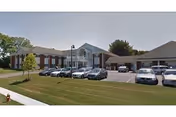 Exterior view of a single-story health and rehabilitation center building with a parking lot in front containing several parked cars. The building has a white facade with brick accents and a peaked roof entrance. There is a grassy area with a small tree near the sidewalk.