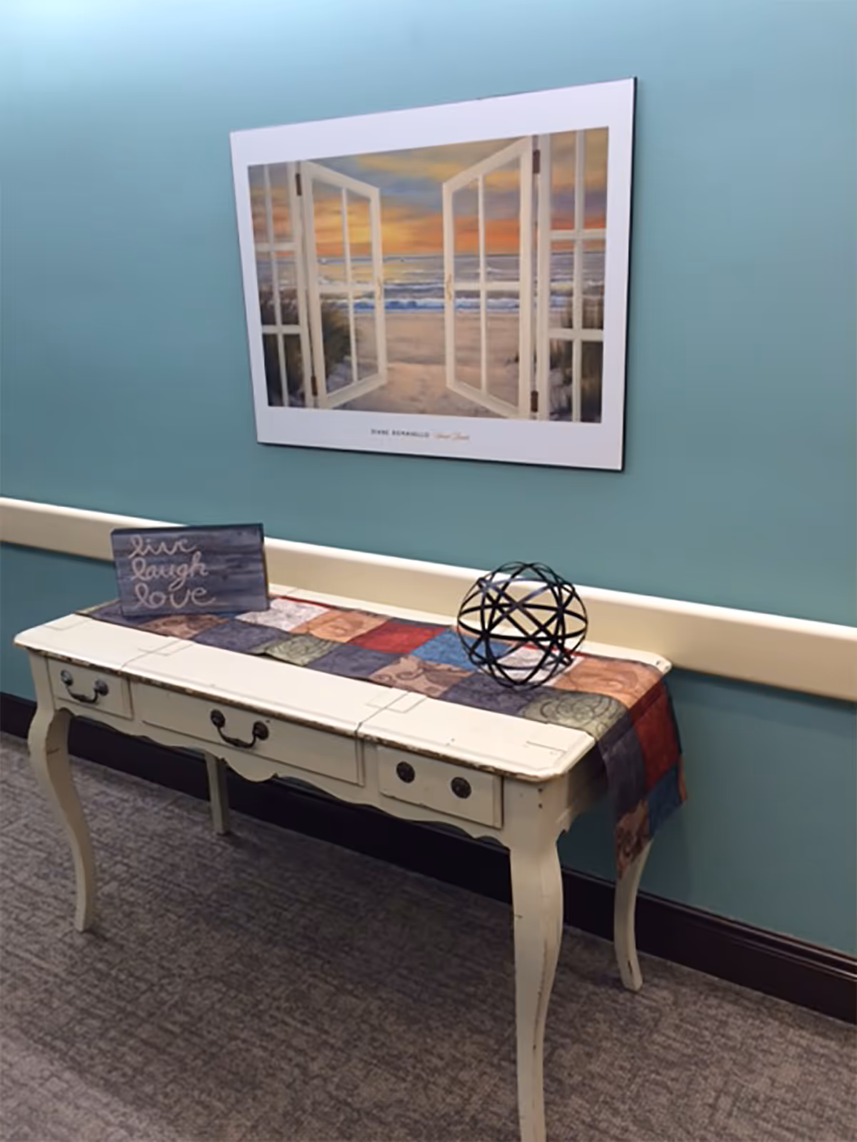 A decorative hallway table with three drawers, painted in a distressed white finish, is placed against a teal wall. On top of the table is a colorful patchwork runner, a small sign with the words 'live laugh love,' and a black metal spherical sculpture. Above the table hangs a framed picture depicting an open window view of a beach at sunset.