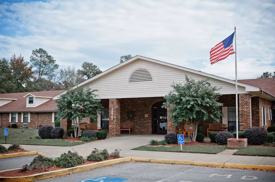 Front entrance of a brick senior care facility with a covered portico, benches, trees, and an American flag.