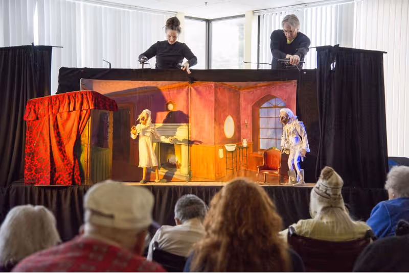 A puppet show is being performed on a small stage with a backdrop depicting a room with a fireplace and window. Two puppeteers dressed in black manipulate puppets dressed in old-fashioned clothing. Several elderly audience members watch the performance.