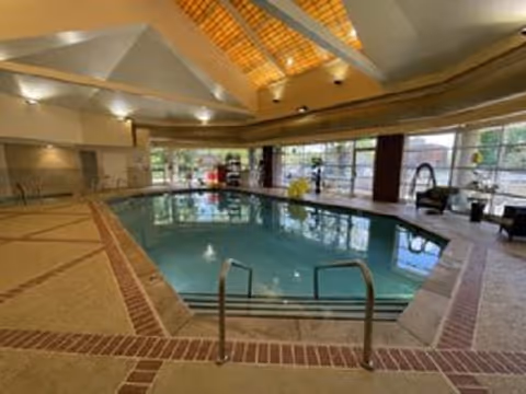 Indoor swimming pool area with large windows allowing natural light, a high ceiling with exposed beams, pool handrails, and seating along the sides.
