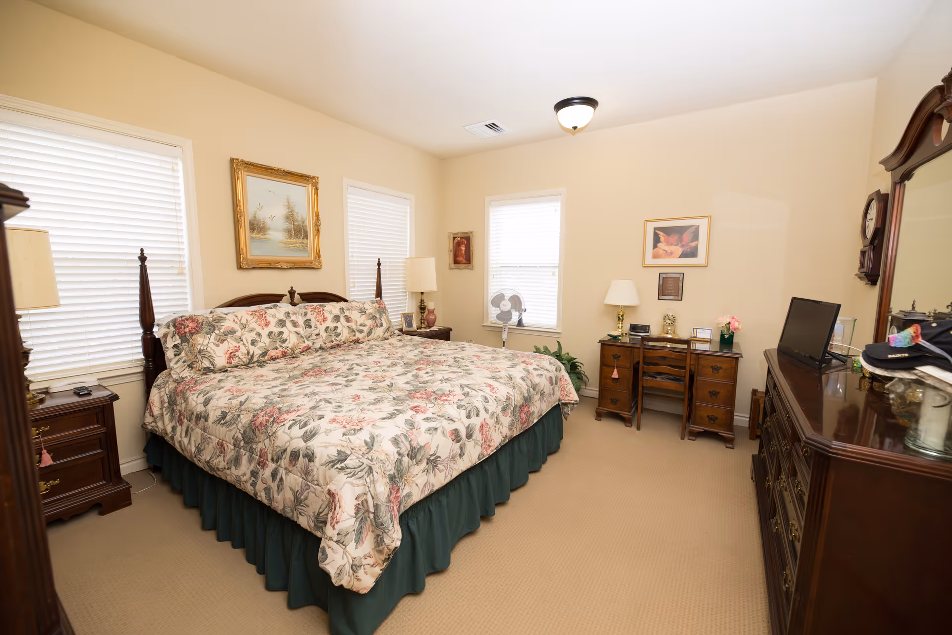 A bedroom with a large bed covered in floral bedding and a green bed skirt. The room has beige walls and carpet, two windows with white blinds, a wooden dresser with a mirror, a wooden desk with a chair, a standing fan, and several framed pictures on the walls. There are two table lamps on either side of the bed and various decorative items on the furniture.