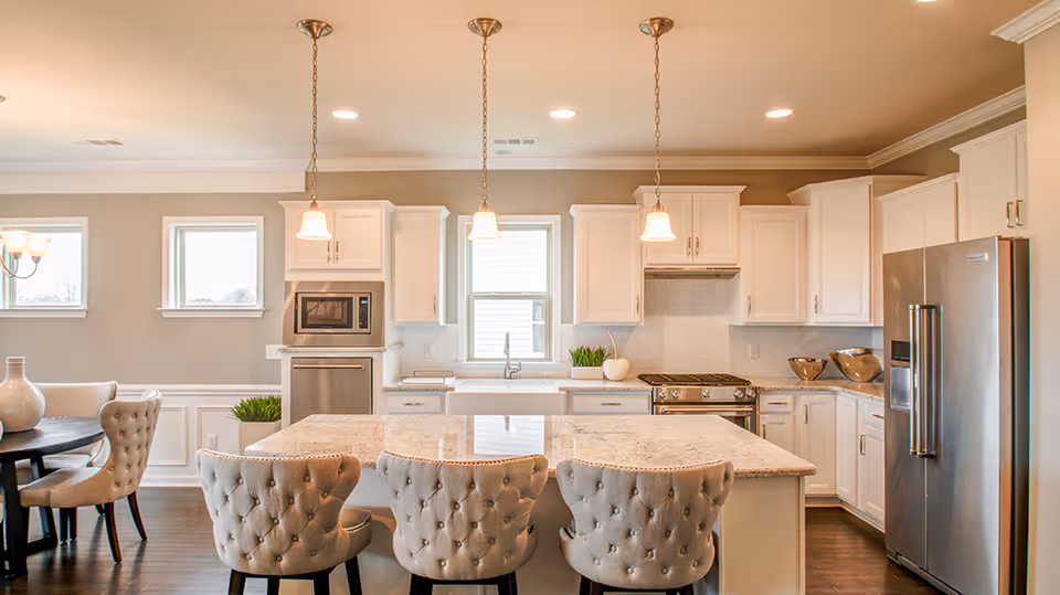 Bright and spacious kitchen with white cabinetry, a large marble island with three tufted chairs, stainless steel appliances including a refrigerator, oven, and microwave. Pendant lights hang above the island, and there is a dining area with a round table and upholstered chairs visible to the left.
