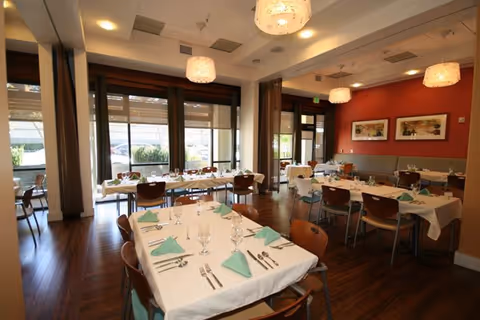 Well-lit dining room with multiple tables set with white tablecloths, folded green napkins, and place settings.