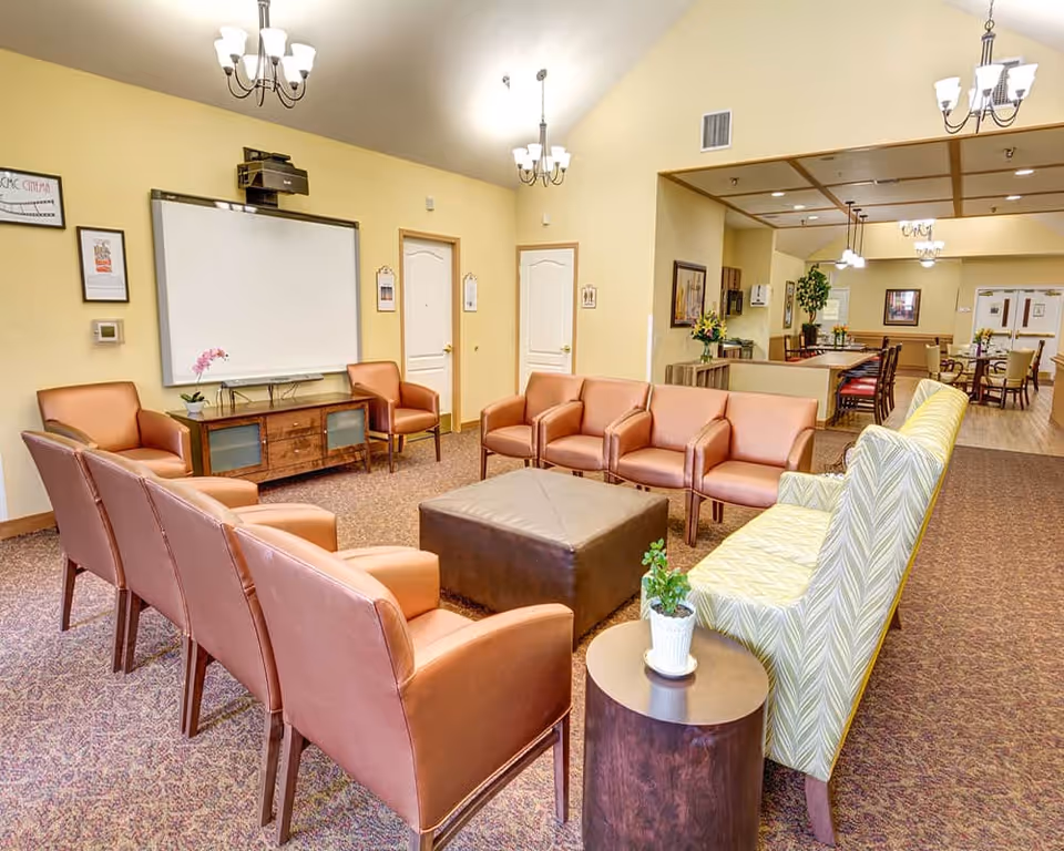 Bright common lounge with chairs and sofas arranged around a central ottoman, a wall-mounted screen, and a dining area visible in the background.