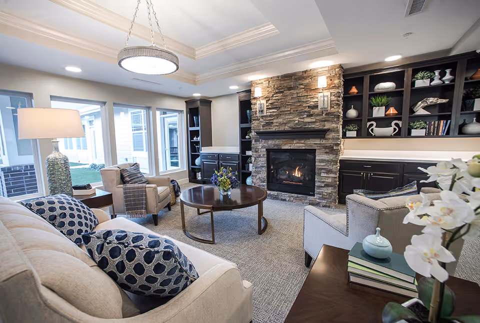 A cozy living room with a beige sofa adorned with blue patterned pillows, a round wooden coffee table with a flower arrangement, two armchairs, a stone fireplace with a fire burning, built-in dark wood shelves with decorative items, and large windows letting in natural light.