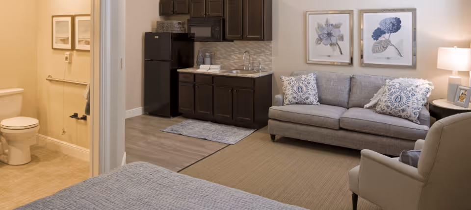 Interior view of a senior living facility room showing a small kitchenette with dark cabinets, a black refrigerator, and a sink. Adjacent to the kitchenette is a living area with a gray sofa adorned with patterned pillows, a side table with a lamp and picture frame, and a comfortable armchair. To the left, a bathroom with a toilet and grab bars is visible through an open door. The room has neutral-colored walls and flooring, with framed floral artwork hanging above the sofa.