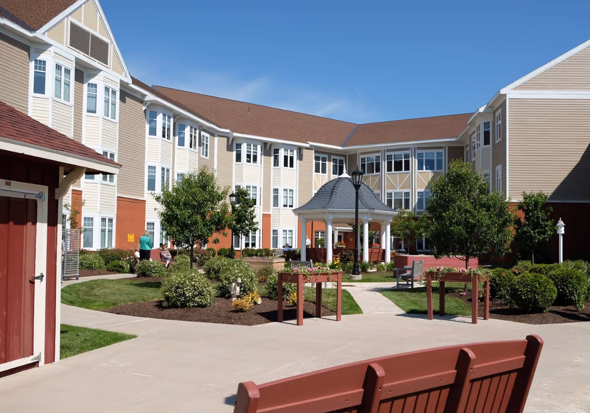 Courtyard with a gazebo, pathways, benches and landscaping in front of a multi-story senior living building under a blue sky.
