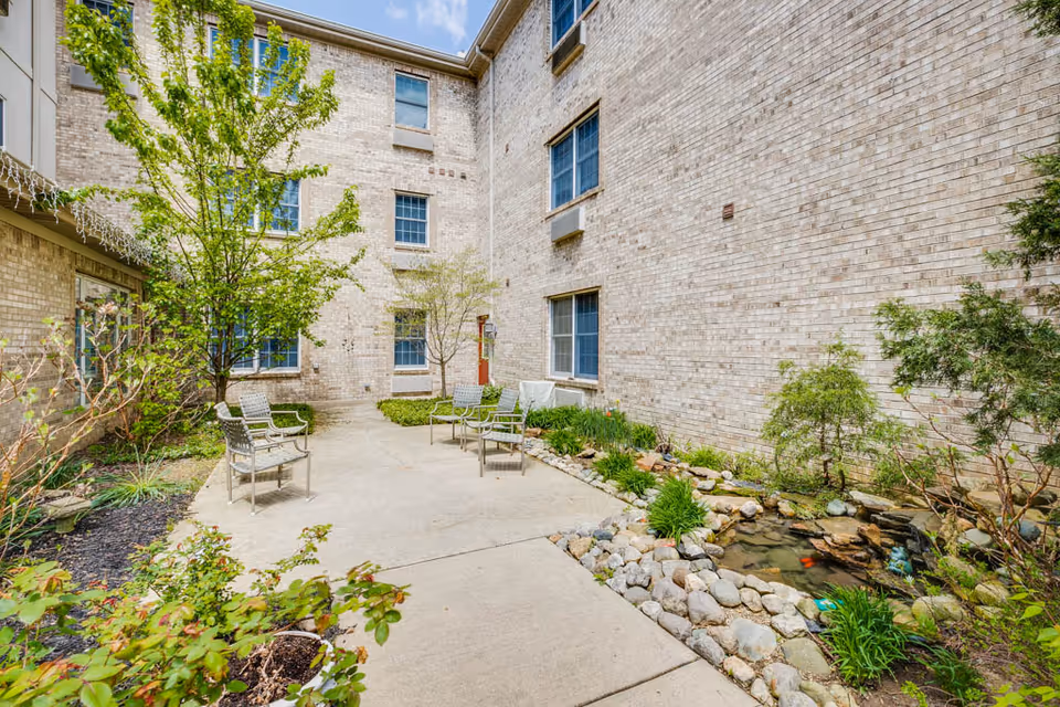 Outdoor courtyard area at Fairborn Assisted Living featuring a concrete pathway, several metal chairs, small trees, shrubs, and a decorative rock-lined pond with water and plants, surrounded by a light brick building under a partly cloudy sky.
