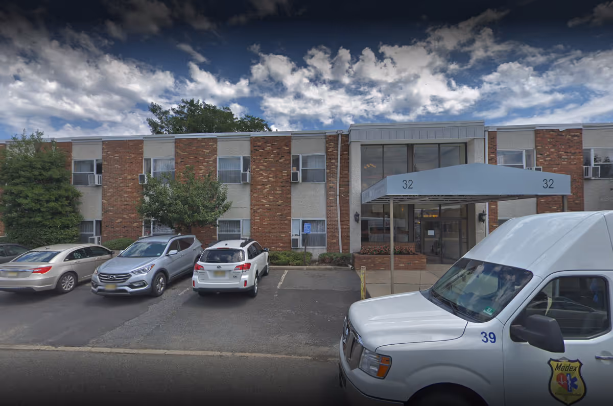 Front exterior view of a two-story brick and concrete building with a covered entrance marked with the number 32. Several cars are parked in front, including a white Medex transportation van. The sky is partly cloudy.