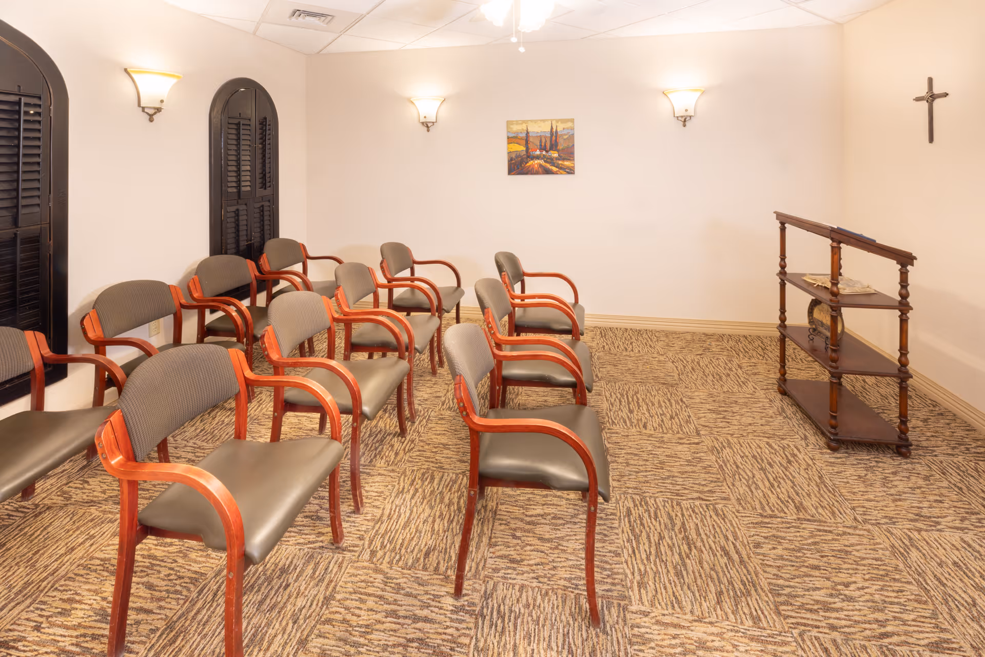 Small interior meeting/chapel room with rows of wooden-framed chairs facing a lectern and a cross on the wall.