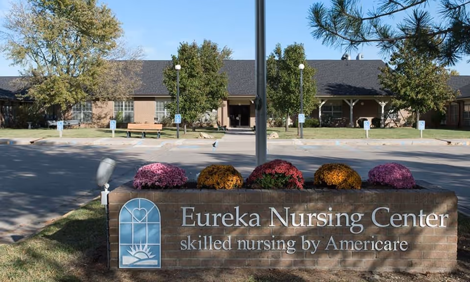 Exterior view of Eureka Nursing Center building with a brick sign in front displaying the facility name and slogan. The sign is decorated with colorful flowers, and there are trees and a parking area with handicap parking signs visible.