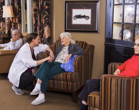 A caregiver kneels and holds hands with an elderly woman seated in a striped armchair in a cozy living room setting. Two other elderly women are seated nearby in similar armchairs. The room features floral curtains, a framed picture of a vintage car on the wall, and a glass-front cabinet with decorative items.