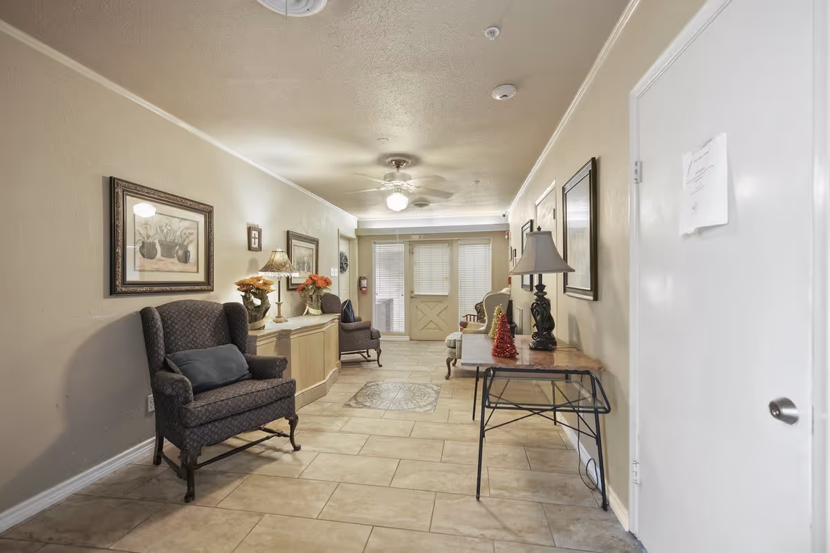 A hallway in a senior living facility with beige walls and tiled floor. The hallway is furnished with armchairs, a sideboard with flower arrangements and lamps, a table with a lamp and small decorative Christmas trees. There are framed pictures on the walls and a ceiling fan with light. At the end of the hallway, there are glass doors with blinds.