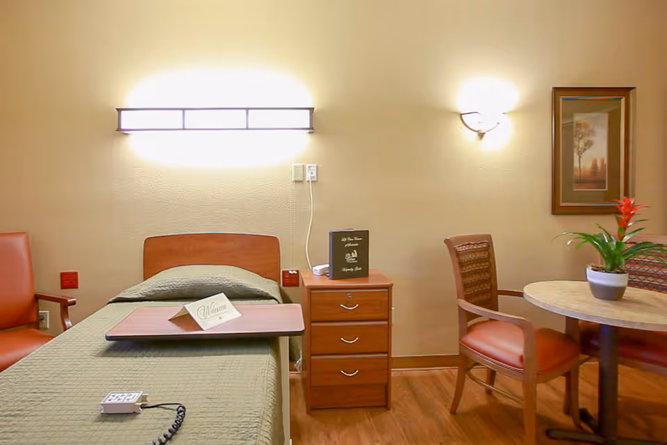 A cozy senior living room with a single bed covered in a green quilt, a wooden bedside table with drawers, a small table with a welcome card on it, two wooden chairs with orange cushions, a round table with a potted plant, and a framed picture on the wall. The room is softly lit by two wall-mounted lights.