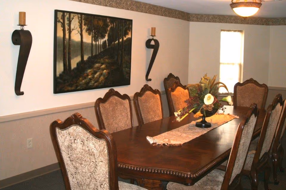 A formal dining room with a long wooden table surrounded by eight upholstered chairs. The table has a decorative runner and a floral centerpiece. On the wall, there is a large painting of a forest scene and two wall-mounted candle holders. A window with light filtering through is visible in the background.