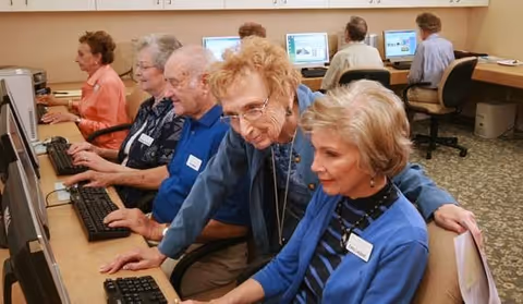 A group of elderly people seated in a computer lab, using desktop computers. One woman is standing and assisting another woman who is seated. The room has beige walls and carpeted flooring.