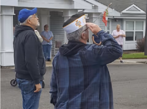Two elderly men standing outside a building, one saluting while wearing a veterans' cap, and the other looking up wearing a blue baseball cap. In the background, two women stand near the building entrance.
