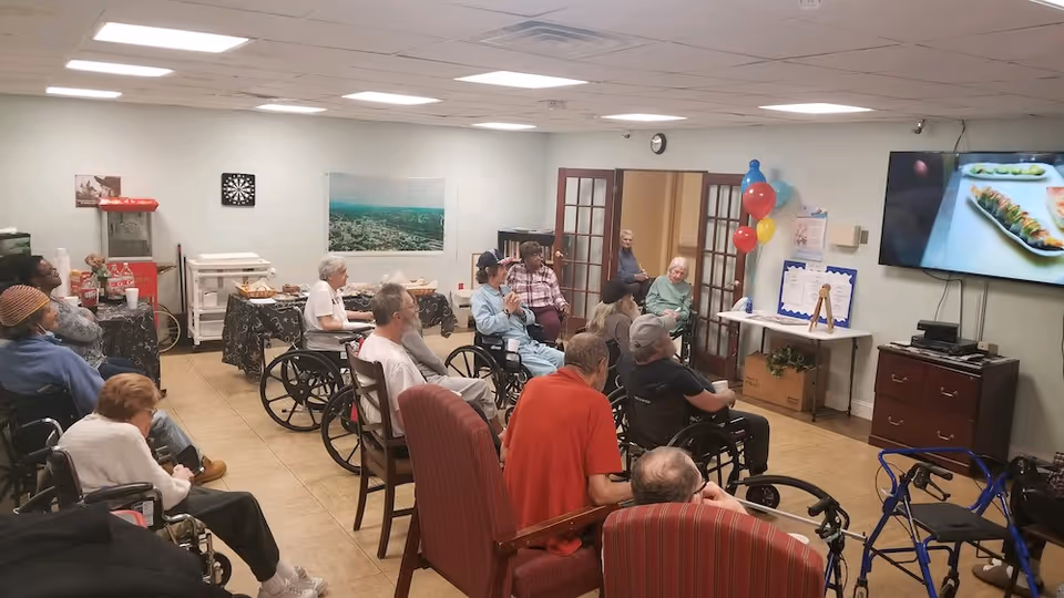 A group of elderly residents in wheelchairs seated in a communal activity room watching a television.