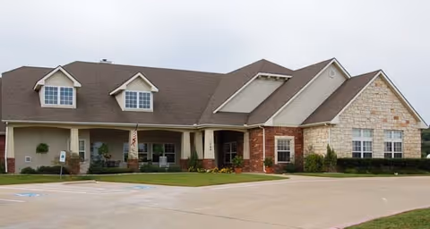 Exterior view of a single-story senior living facility building with a combination of brick and stone facade, multiple windows, and a covered entrance. The building is surrounded by a paved parking area and some landscaping with grass and small bushes.