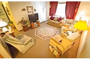 A cozy living room with beige carpet, a round patterned rug in the center, and several upholstered chairs and sofas arranged around a flat-screen TV on a wooden stand. The room has red curtains on the window, a floor lamp, a table lamp, and decorative items including framed pictures and a flower arrangement.
