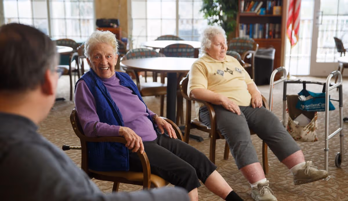 Two elderly women sitting on chairs in a common area of a senior living facility, engaging in a seated exercise with their legs extended. A man is partially visible in the foreground. The room has round tables and chairs, carpeted floor, large windows, and a bookshelf with an American flag in the background.