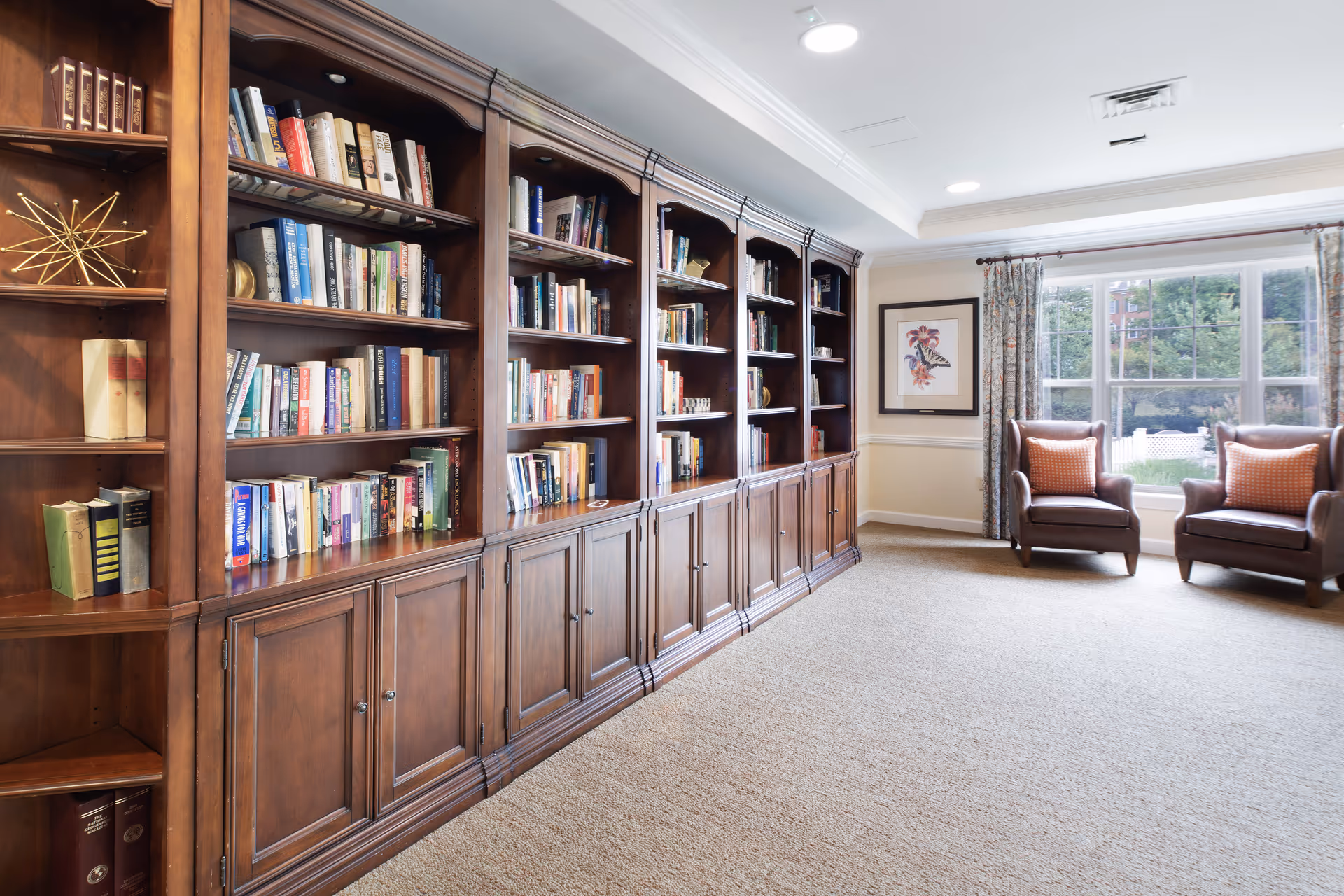 Sunlit reading room with large wooden built-in bookshelves filled with books and two armchairs by a window.