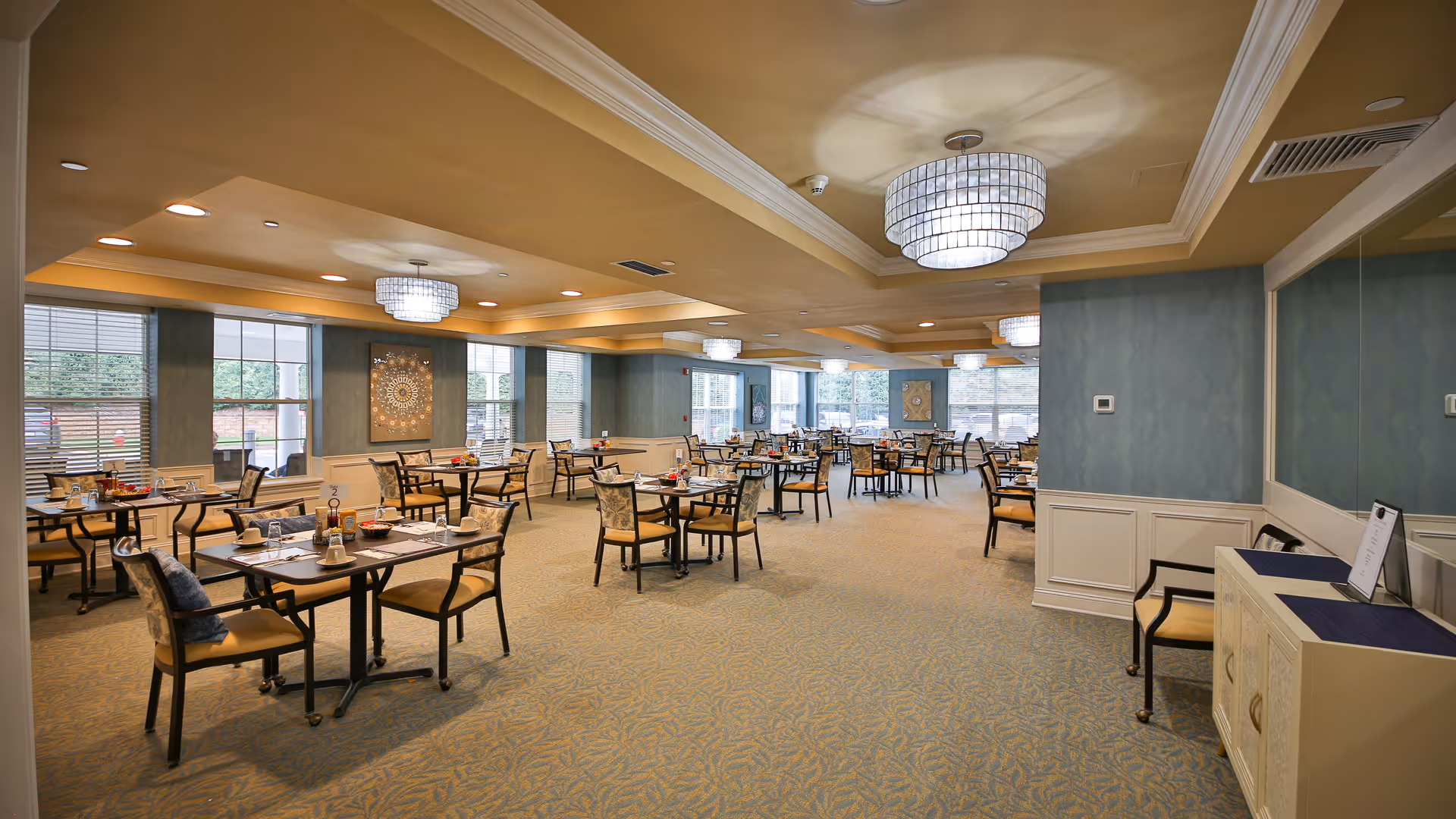 A spacious dining room in Avalon Assisted Living at Bridgewater with multiple tables and chairs arranged neatly. The room features large windows allowing natural light, elegant chandeliers hanging from a tray ceiling, and decorative wall art. The carpeted floor and soft blue walls create a warm and inviting atmosphere.