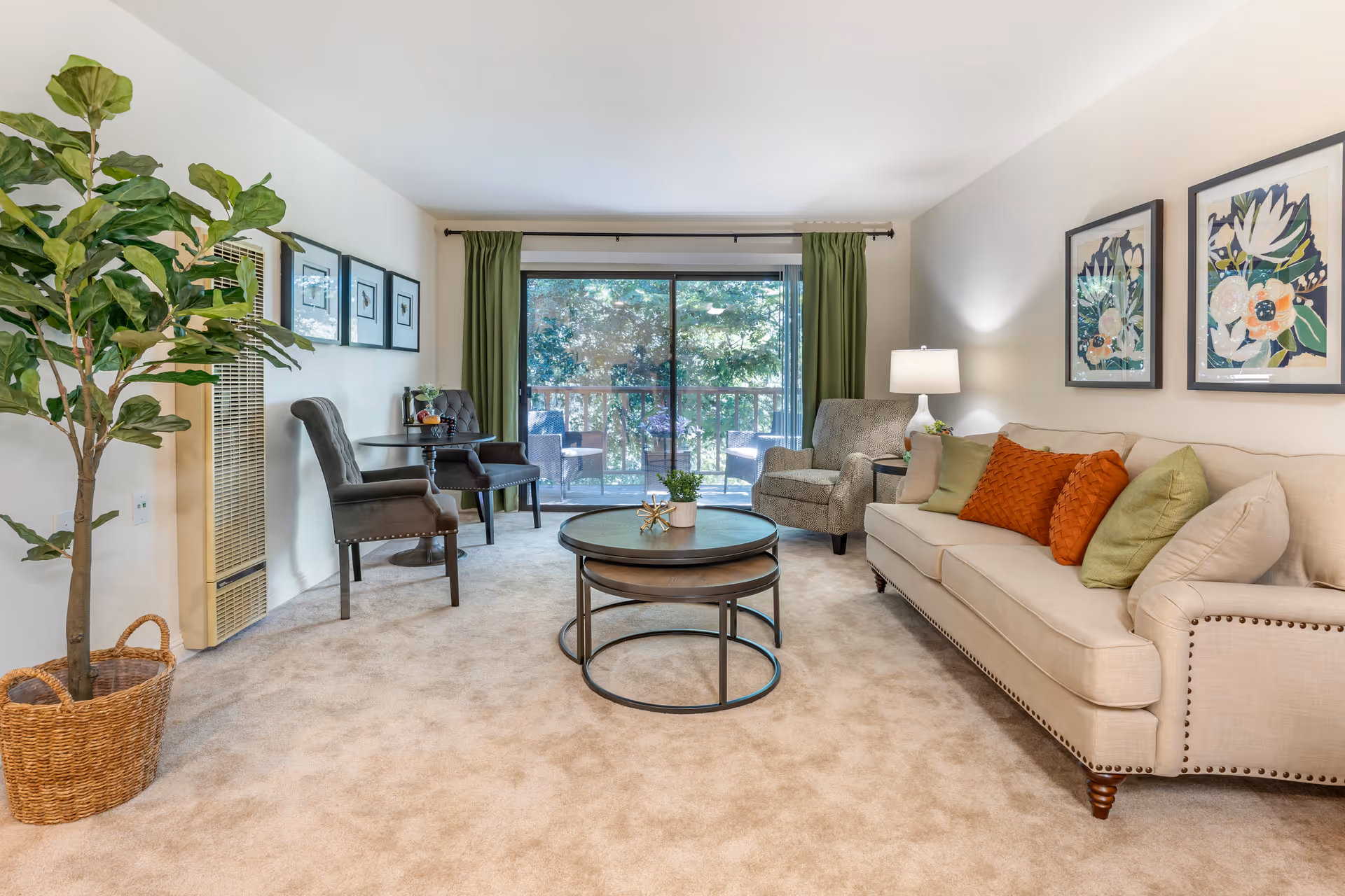 A bright living room with a beige sofa adorned with green and orange pillows, a gray armchair, two dark gray chairs with a small round table between them, a round nested coffee table with a small plant on top, green curtains framing a large sliding glass door leading to an outdoor patio, and a large potted plant in a wicker basket on the left side.