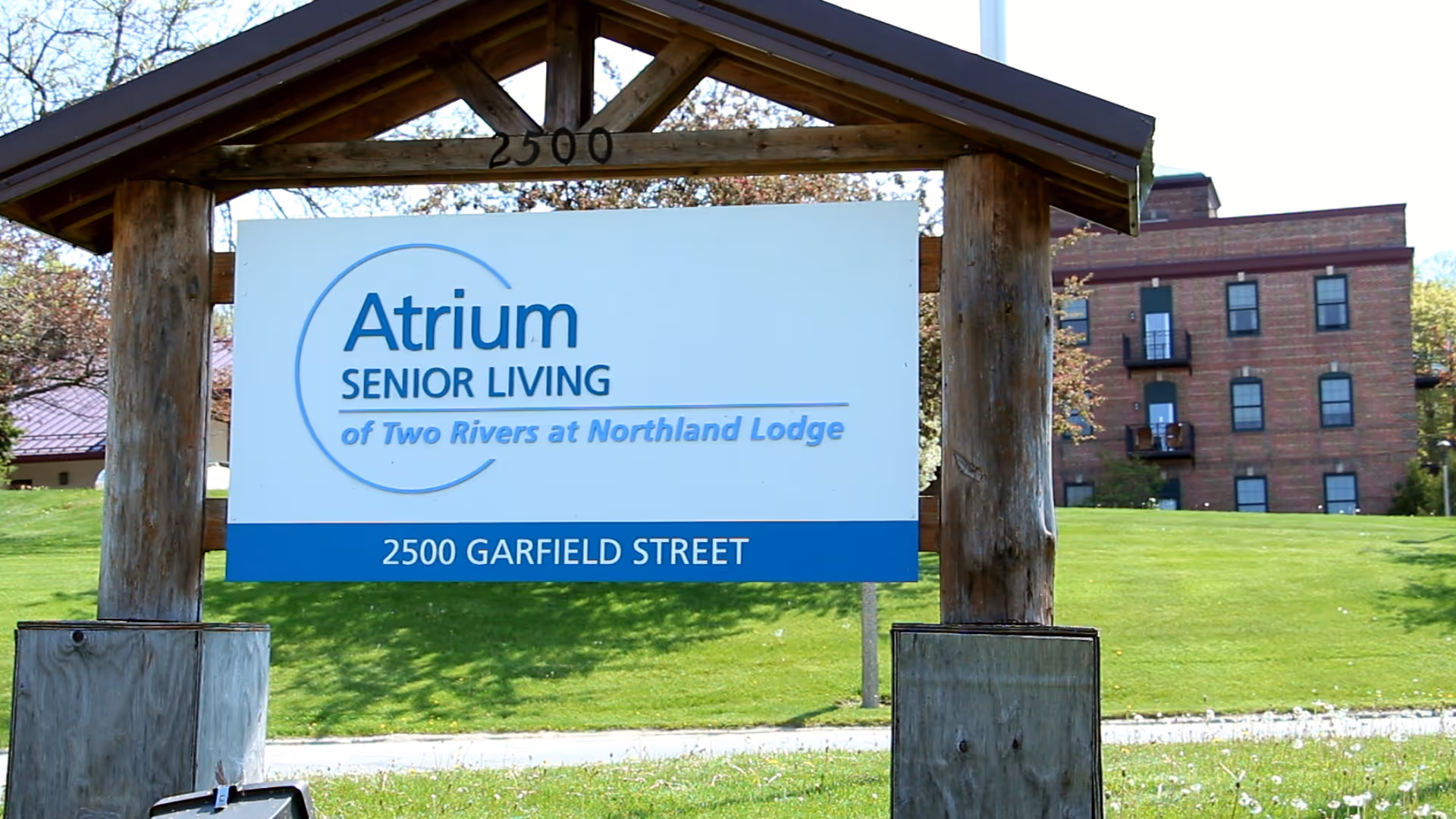 Outdoor view of a wooden sign structure with a white and blue sign that reads 'Atrium Senior Living of Two Rivers at Northland Lodge, 2500 Garfield Street' with a brick building and green lawn in the background.