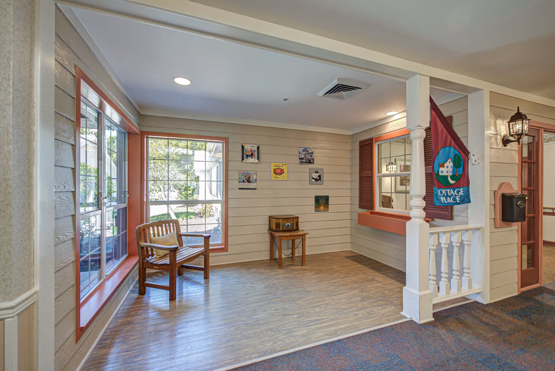 Bright interior seating nook with a wooden bench by large windows, a small side table, and a 'Cottage Place' flag by a service window.