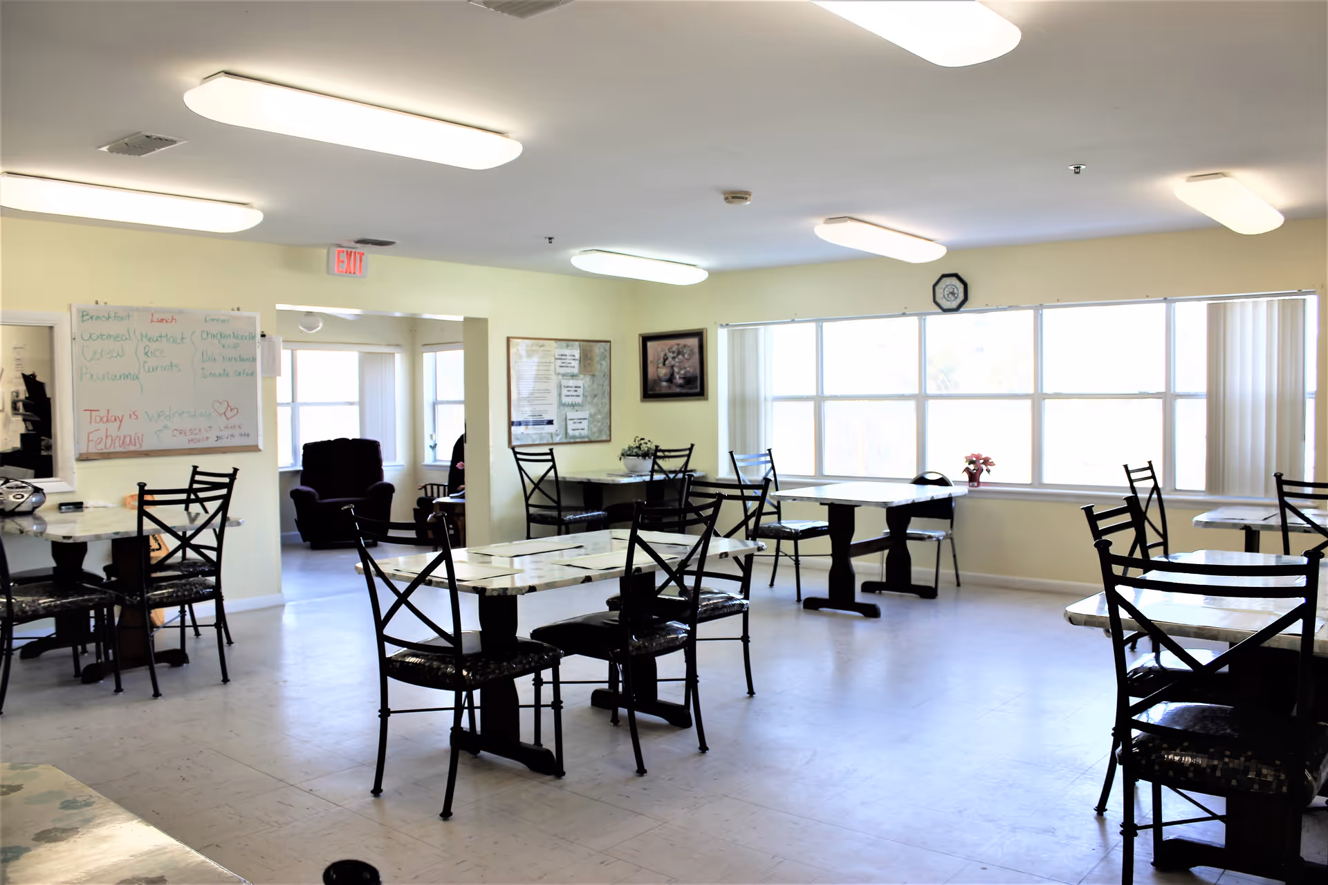 A bright dining room with several tables and chairs arranged neatly. Large windows on one side let in natural light. A whiteboard with daily meal information and a bulletin board are visible on the walls. The room has a clean and simple design with light-colored walls and ceiling lights.