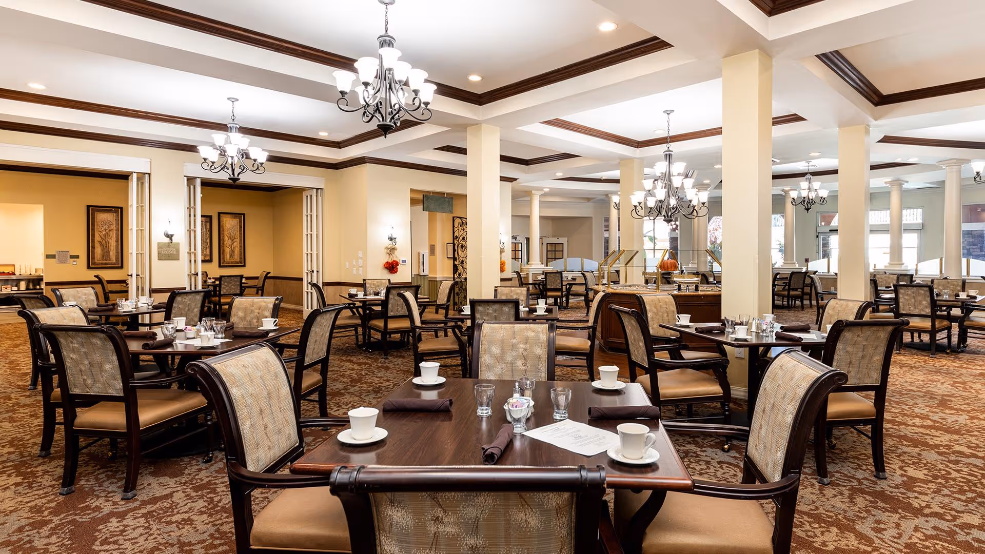 A spacious dining room in a senior living facility with multiple dark wood tables and cushioned chairs arranged neatly. Each table is set with white cups, glasses, and napkins. The room features beige walls, patterned carpet, decorative wall art, and elegant chandeliers hanging from a coffered ceiling.