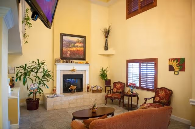 A cozy living room with beige walls and tiled floor featuring a fireplace with a framed painting above it. There are two red upholstered armchairs with floral patterns, a wooden coffee table, a small side table with a plant, and a large potted plant near an archway. A flat-screen TV is mounted high on the wall, and a window with wooden blinds lets in natural light.