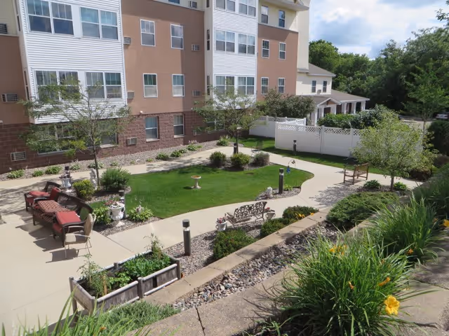 A landscaped courtyard with a grassy lawn, winding walkways, benches, planters and patio seating next to a multi-story residential building.