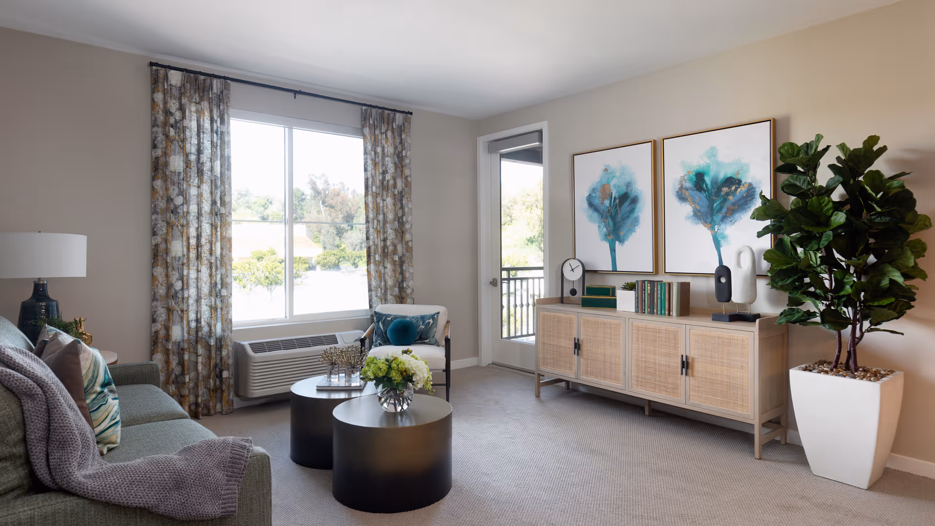 A bright and cozy living room with a green sofa adorned with pillows and a gray throw blanket. In front of the sofa are two round black coffee tables with decorative items including a vase of flowers. A large window with patterned curtains lets in natural light. On the opposite wall, there is a wooden sideboard with books, decorative objects, and two framed abstract paintings of blue trees. A tall potted plant stands next to the sideboard. The room has beige walls and carpeted flooring.